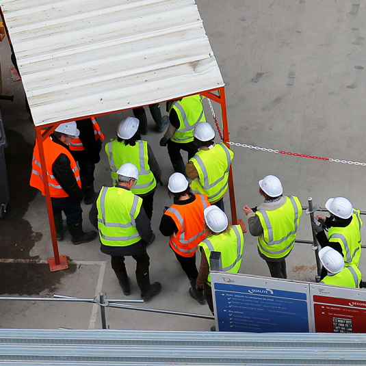 Time tracking software in construction: With the Time App, you have all site accesses under control. Construction workers with yellow and orange high-visibility vests and white helmets enter the construction site.