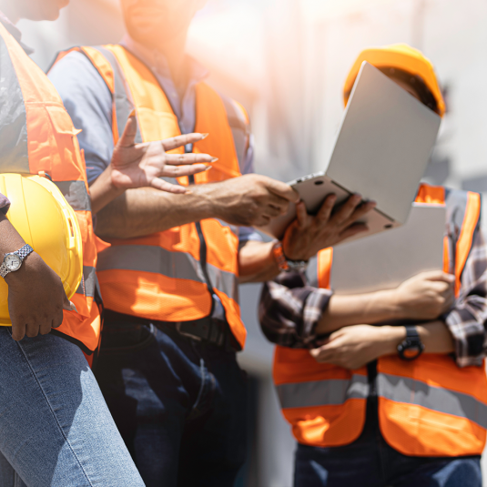 Material-Tracking-Software-Einführung: Binden Sie die User ein. Drei Arbeiter auf der Baustelle unterhalten sich miteinander, sie schauen auf einen Laptop.